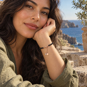 A woman rests her hand near her cheek with a rose gold vermeil and black bead bracelet visible against a seaside terrace.