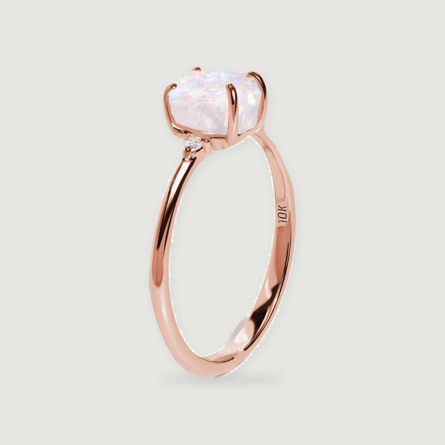A rose gold ring stands upright with a pale moonstone and a visible diamond beside the setting.