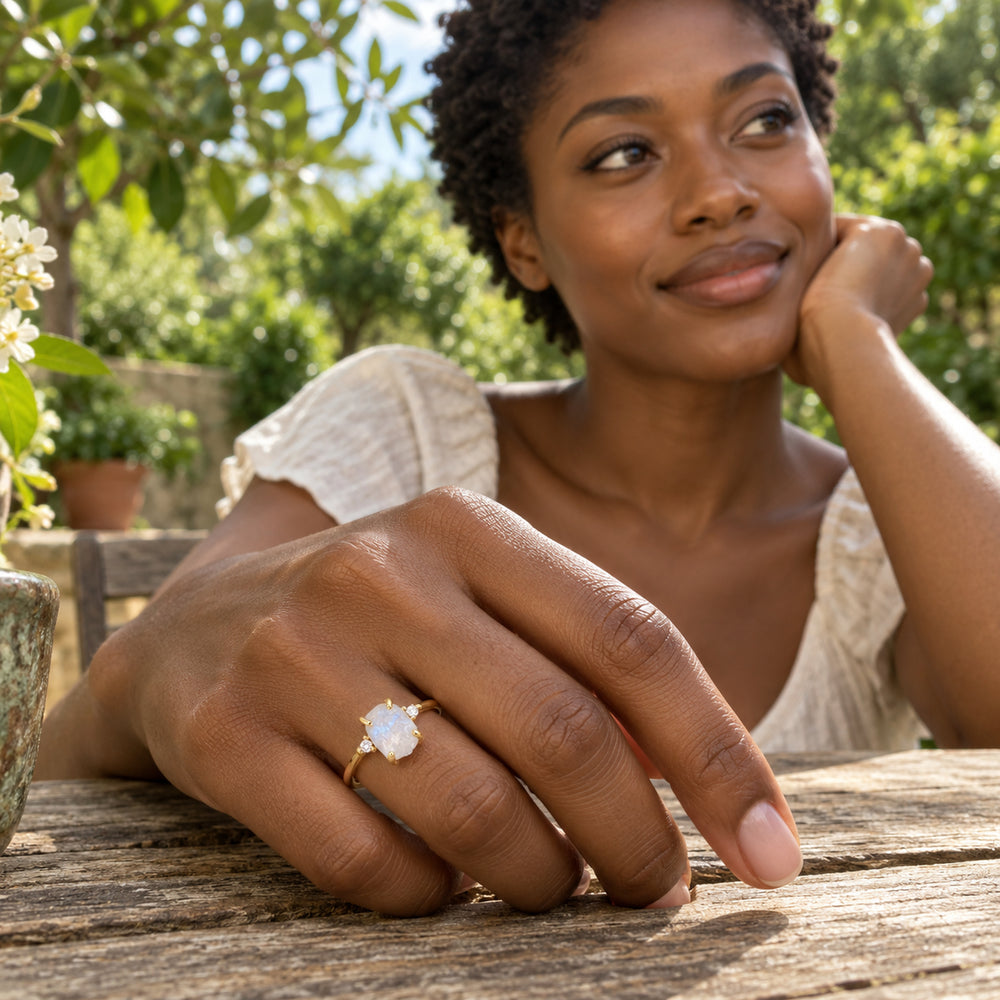 A yellow gold moonstone ring is worn on a hand resting on a wooden table in a leafy garden.