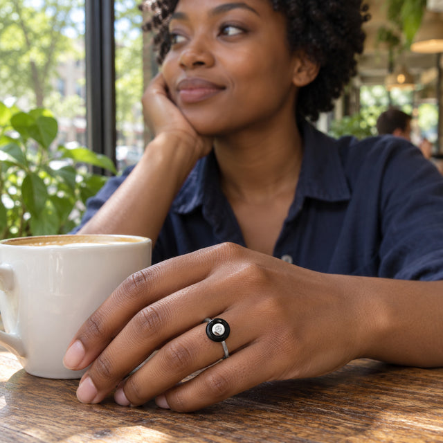 A silver ring with a black circular stone is worn on a hand beside a coffee cup at a cafe window table.