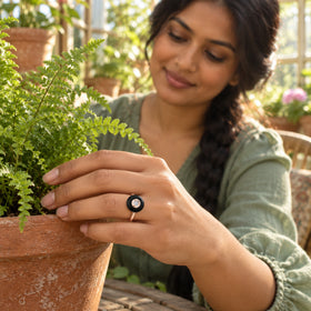 A rose gold ring with a glossy black circular stone is shown on a hand resting against a terracotta planter beside a fern.