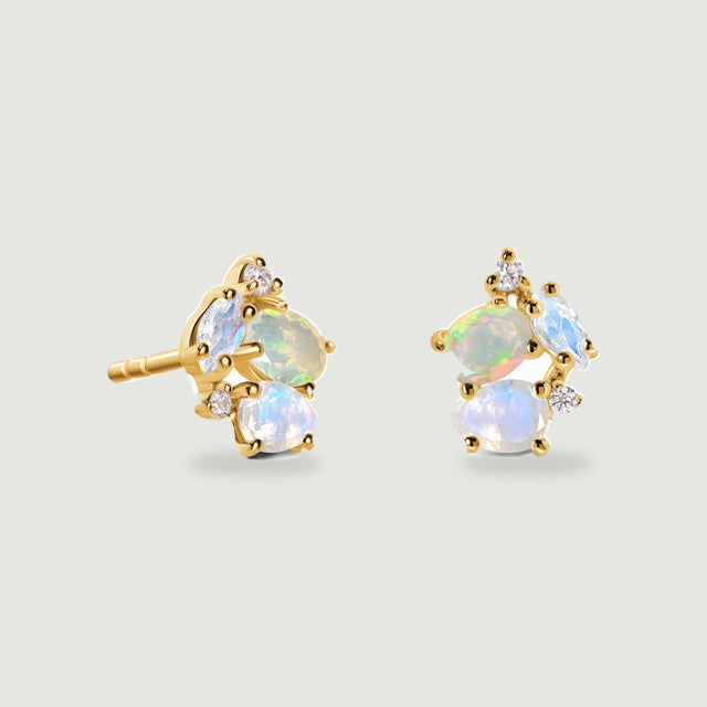 Two yellow gold vermeil stud earrings show an iridescent opal, pale blue moonstones, and small clear accent stones on a light gray background.