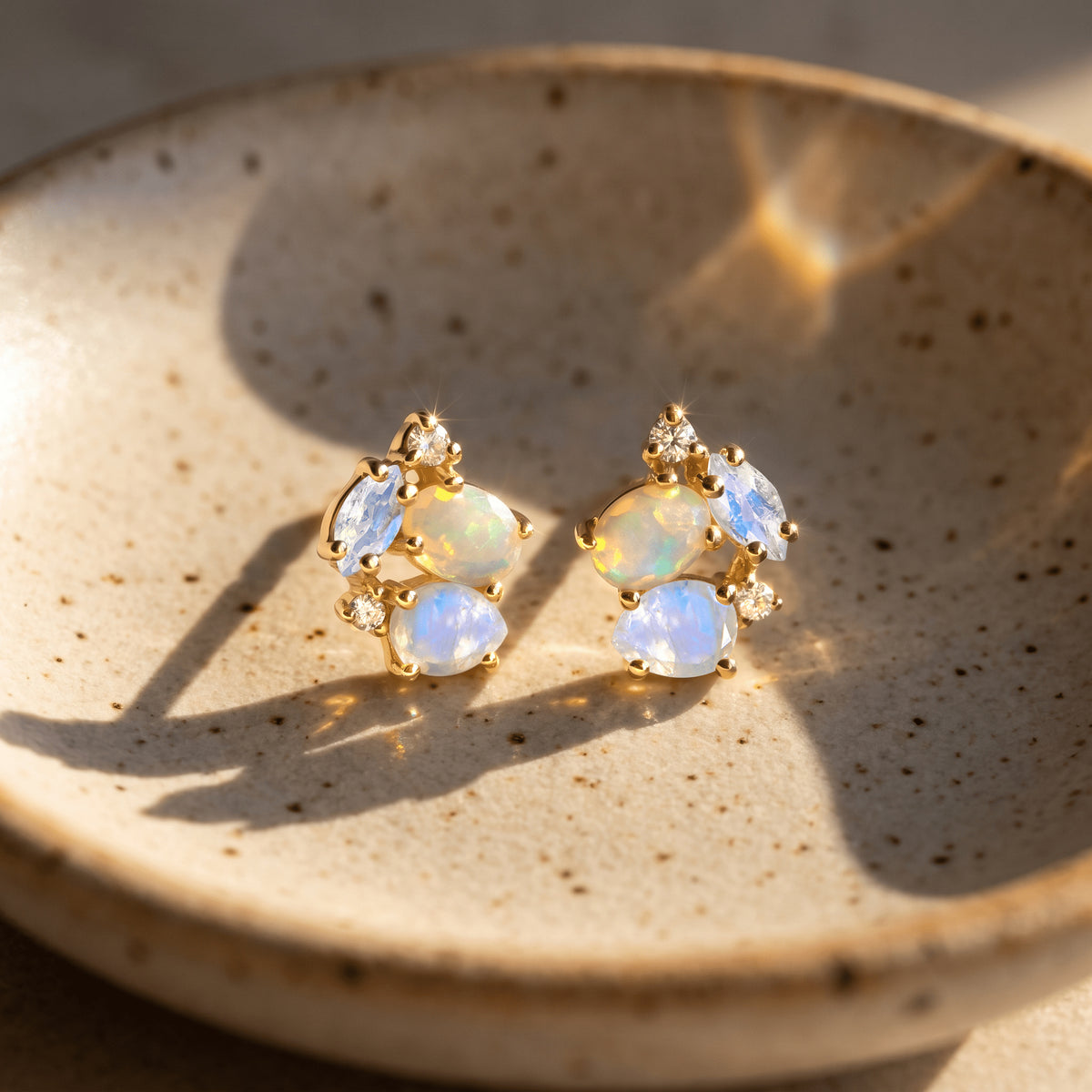 Two yellow gold gemstone studs lie on a speckled stone dish in sunlight with long soft shadows.