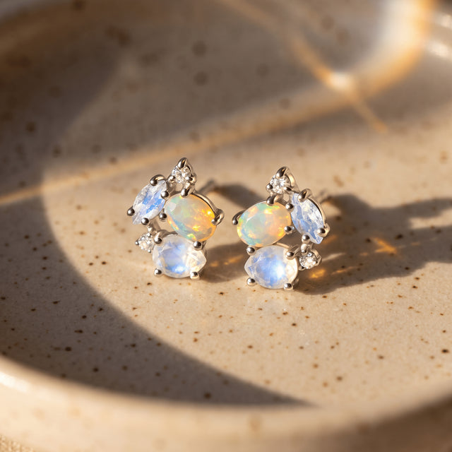 Silver gemstone earrings sit side by side on a warm stone surface, with blue moonstone facets and bright opal reflections.
