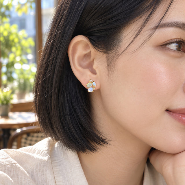 A woman with a black bob wears a yellow gold gemstone cluster stud beside a bright cafe window.