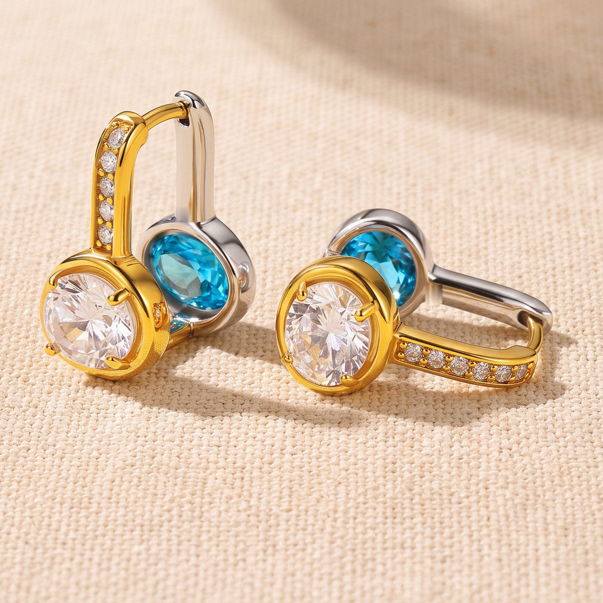 Two reversible hoop earrings rest on textured cream fabric with their clear and blue gemstone faces visible.