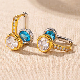 Two reversible hoop earrings rest on textured cream fabric with their clear and blue gemstone faces visible.