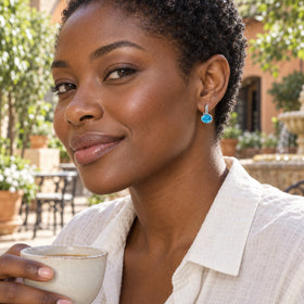 A woman with short natural curls wears a silver-colored hoop with a blue oval gemstone while holding a ceramic cup in a sunny courtyard.