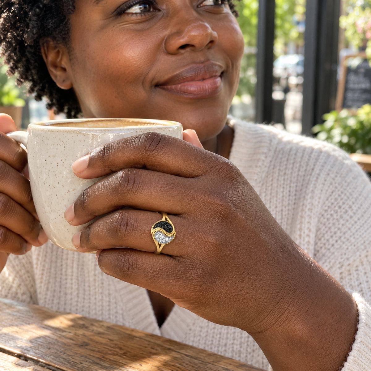 A woman wearing a yellow gold ring holds a speckled ceramic cup beside a sunlit cafe window.