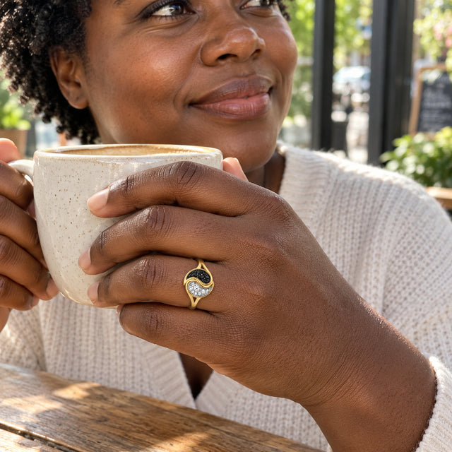 A woman wearing a yellow gold ring holds a speckled ceramic cup beside a sunlit cafe window.