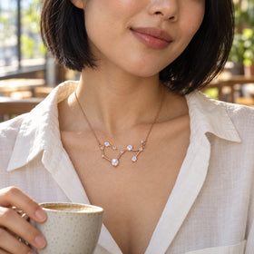 A woman in an ivory blouse wears the rose gold constellation necklace while holding a cup beside a cafe window.