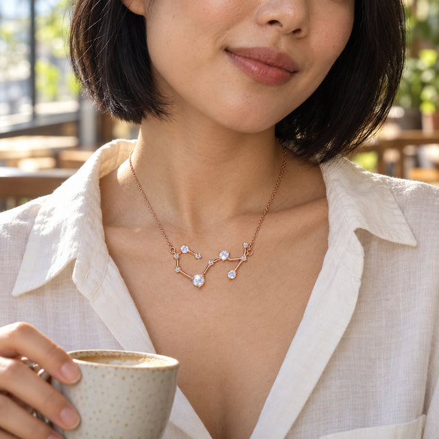 A woman in an ivory blouse wears the rose gold constellation necklace while holding a cup beside a cafe window.