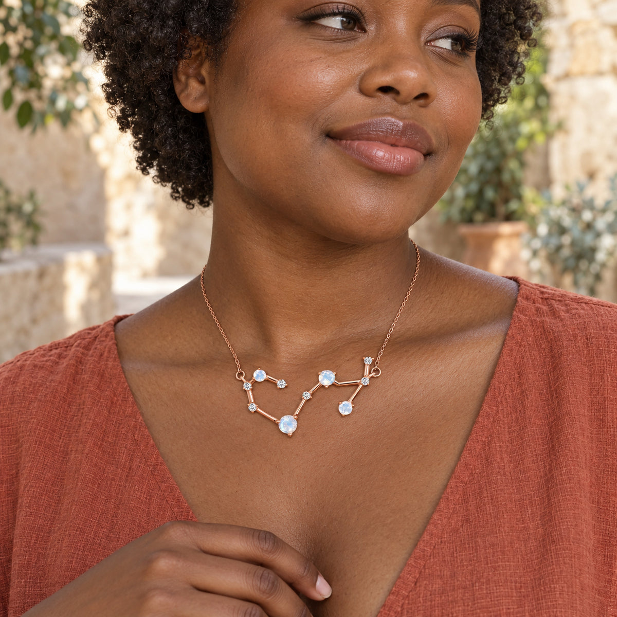 The rose gold necklace rests below a woman's collarbones above a terracotta blouse in a leafy stone courtyard.