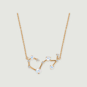 A yellow gold necklace with a curved constellation pendant and round iridescent stones rests on a light gray background.