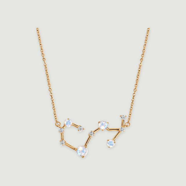 A yellow gold necklace with a curved constellation pendant and round iridescent stones rests on a light gray background.