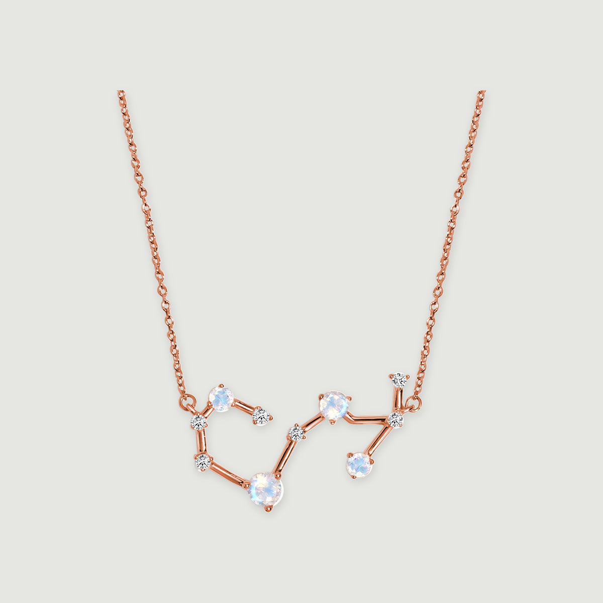 A rose gold chain connects to an open constellation pendant with pale round stones on a light gray surface.