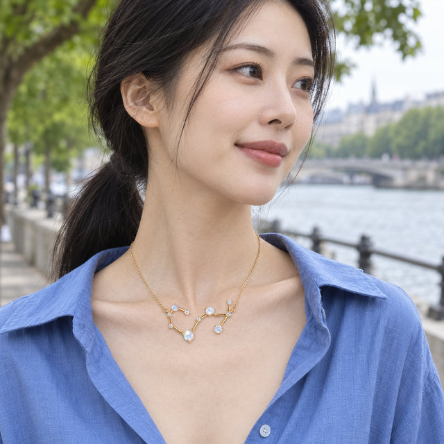 A woman in a white linen shirt wears a yellow gold constellation necklace on a sunlit terrace.