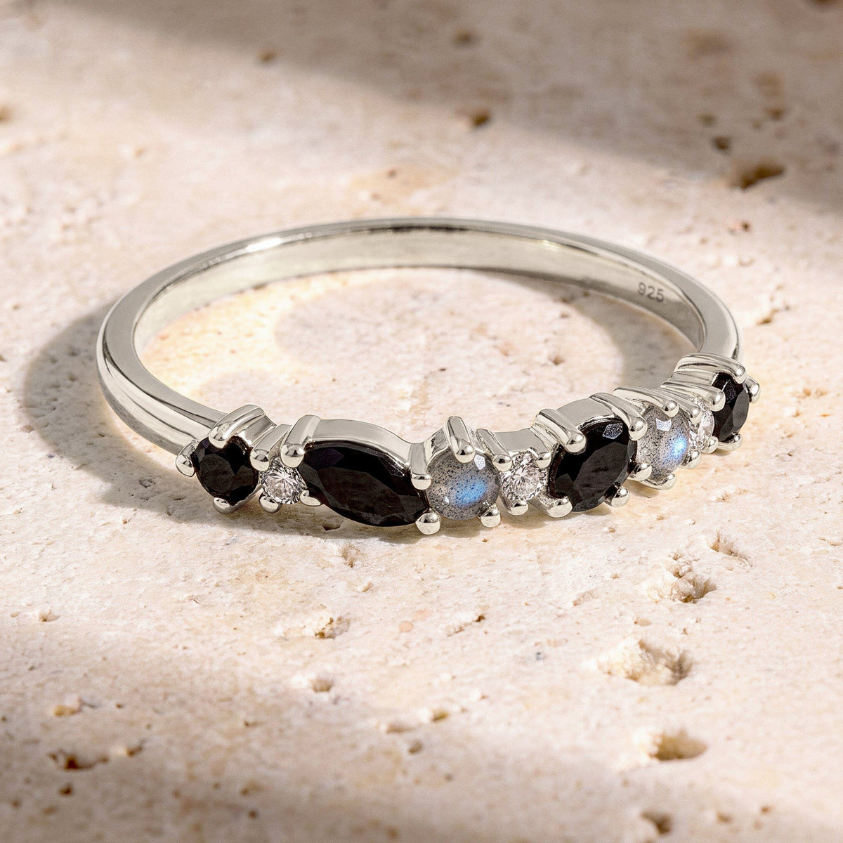 The sterling silver ring lies on a textured cream stone surface with its row of dark and pale stones facing forward.