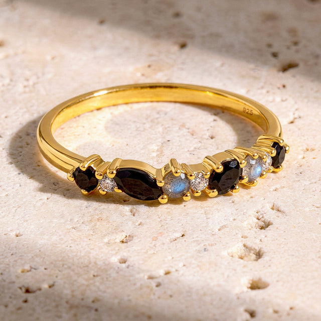 The yellow gold vermeil ring lies on a textured cream stone surface with its row of dark and pale stones facing forward.