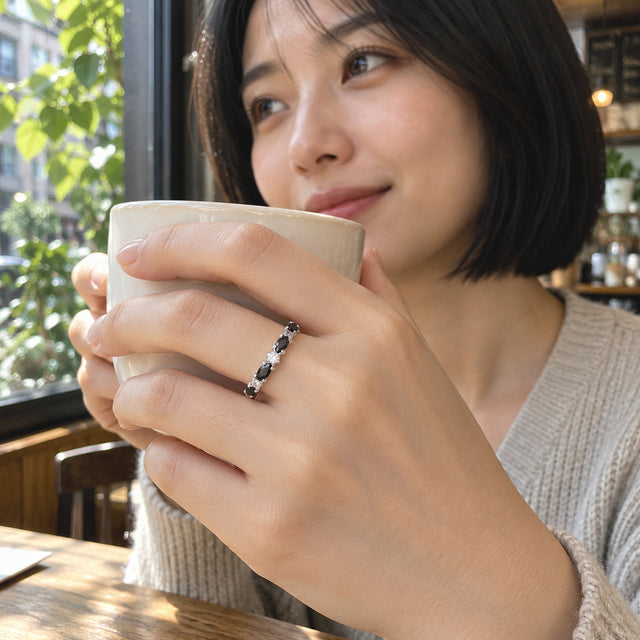 A woman with a black bob holds a cream ceramic cup while wearing the silver ring beside a leafy cafe window.