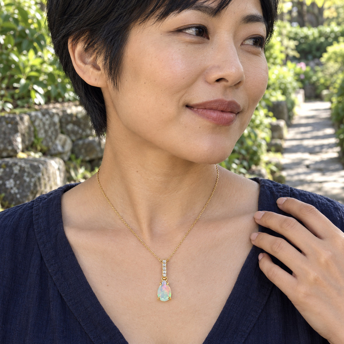 A woman with short black hair wears the yellow gold opal necklace with a navy top on a leafy garden path.