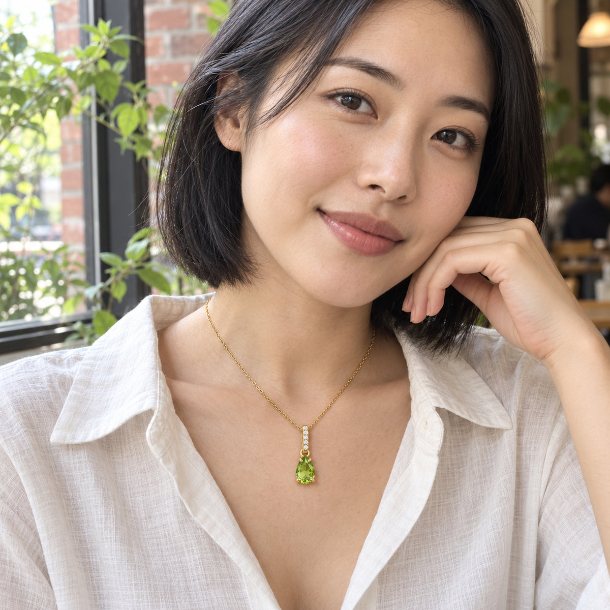 A woman in a white linen blouse wears the yellow gold peridot necklace beside a cafe window with leafy plants.