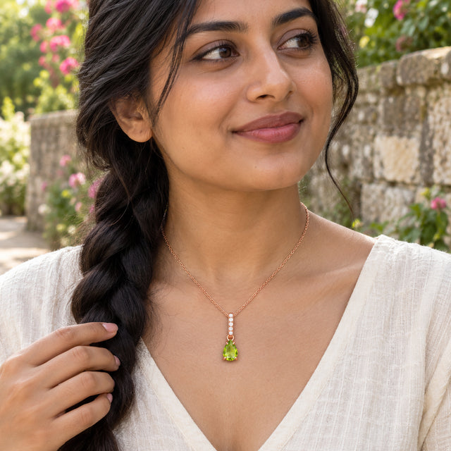 A woman with braided dark hair wears the rose gold peridot necklace with a white top beside a flowering garden wall.
