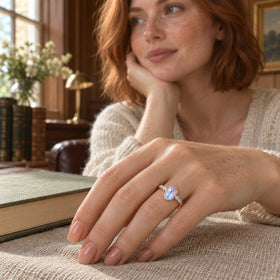 A rose gold moonstone ring is worn on a freckled hand resting on linen beside a closed book.