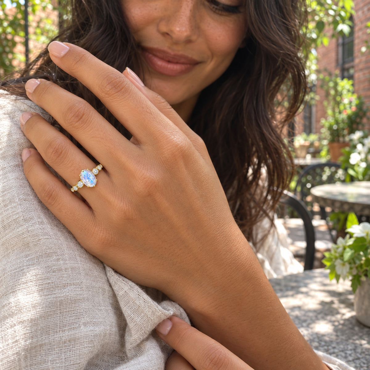 A yellow gold ring with an oval pale blue moonstone is worn on a hand resting against a white linen sleeve in a leafy courtyard.
