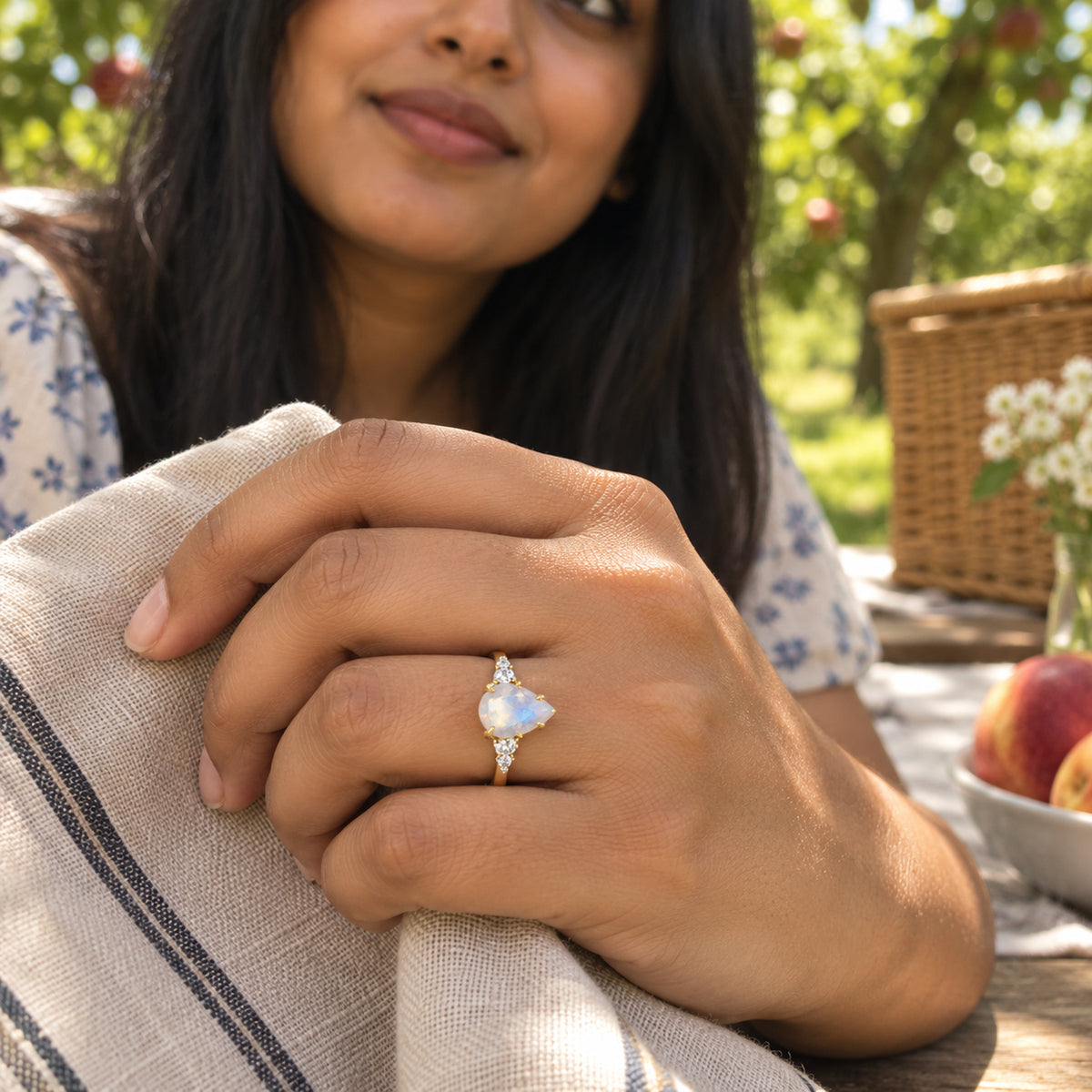 A yellow gold moonstone ring is worn on a hand holding striped linen beside an orchard picnic table.
