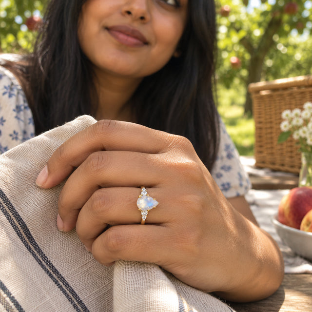 A yellow gold moonstone ring is worn on a hand holding striped linen beside an orchard picnic table.