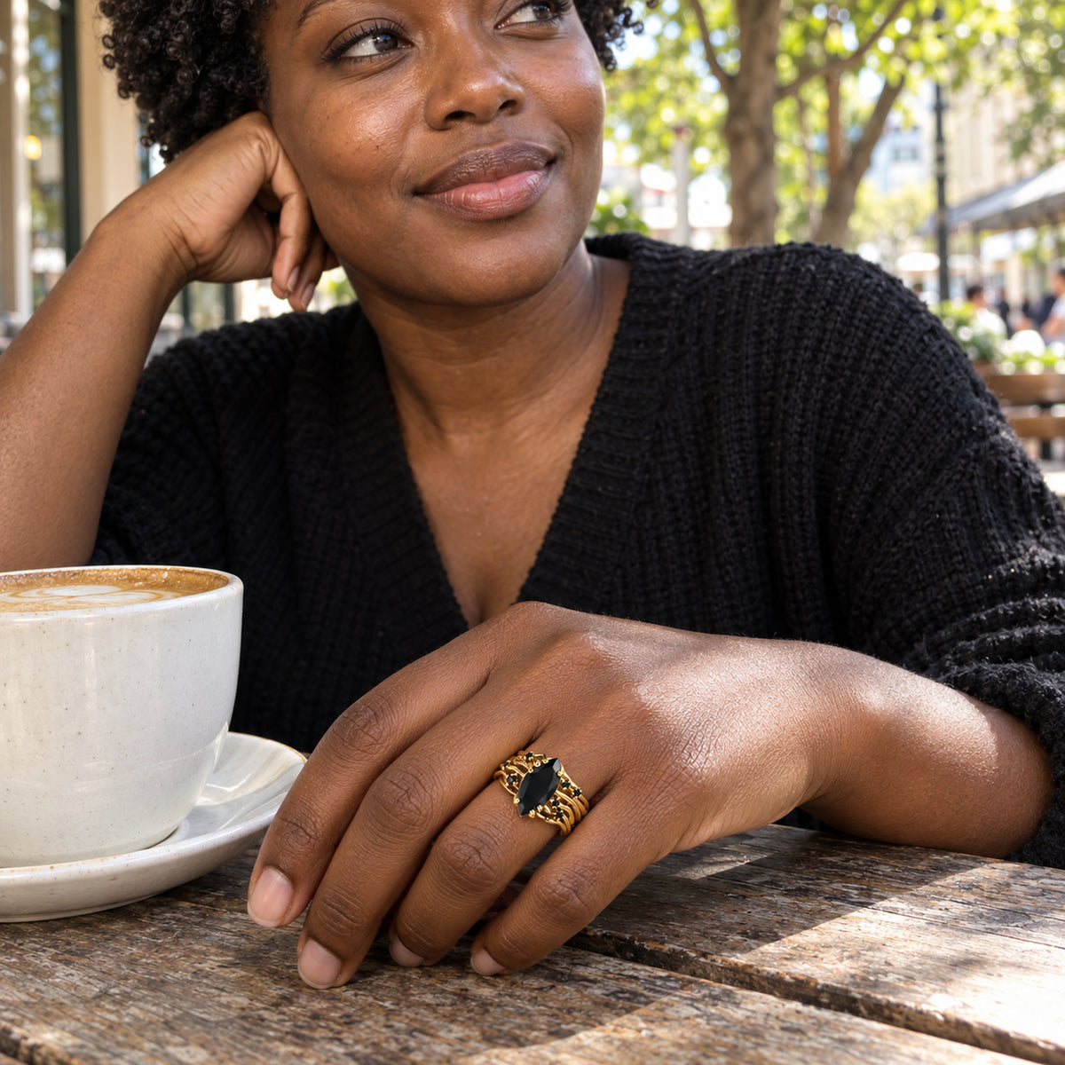 A yellow gold ring with a pointed black stone is worn on a hand beside a white coffee cup at an outdoor cafe.