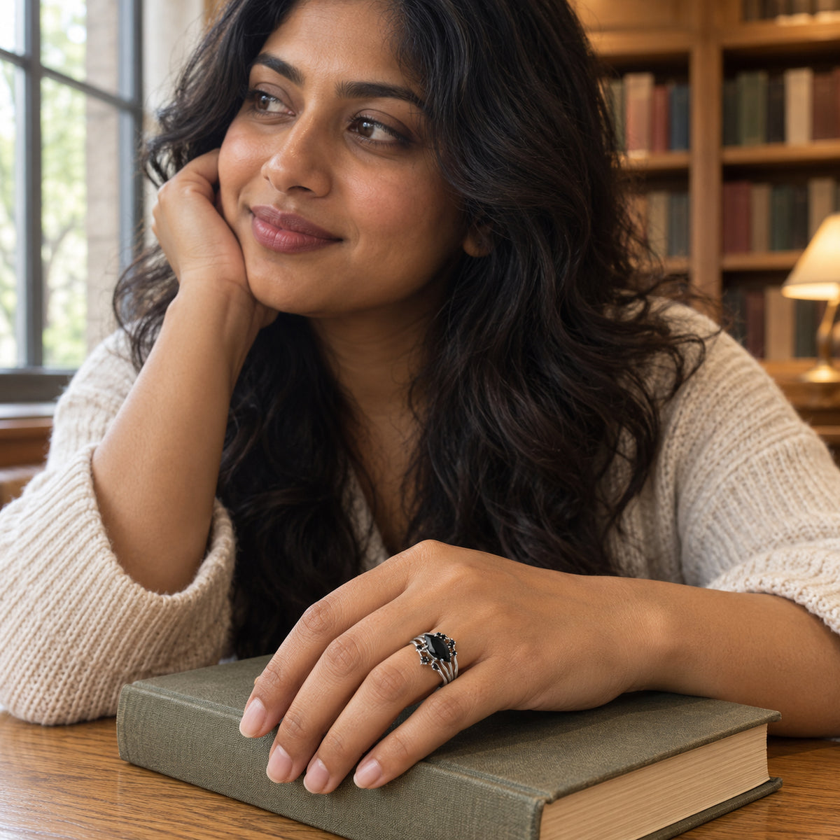 A silver ring with a marquise black stone is worn on a hand resting on a closed book beside a library window.