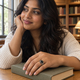 A silver ring with a marquise black stone is worn on a hand resting on a closed book beside a library window.