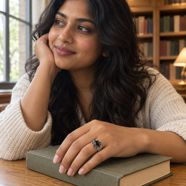 A silver ring with a marquise black stone is worn on a hand resting on a closed book beside a library window.