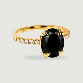 A yellow gold ring with an oval black center stone and clear shoulder stones rests on a pale gray surface.