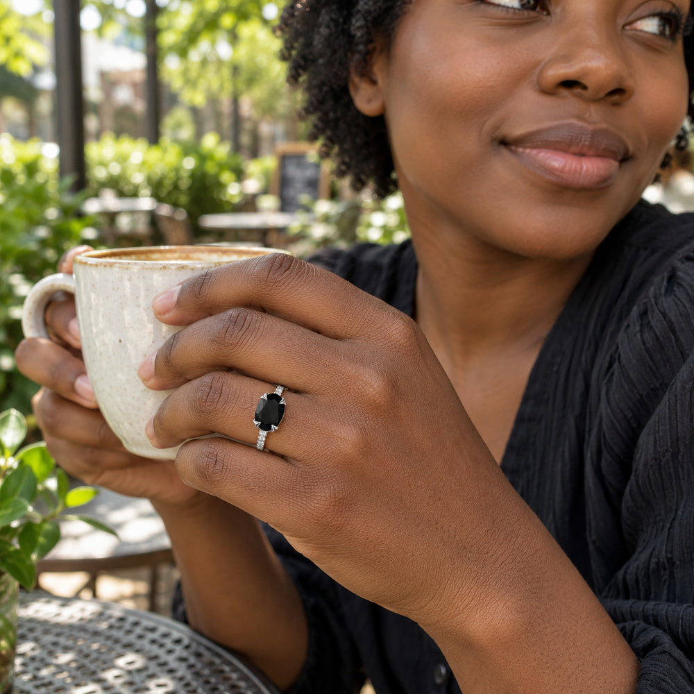 A woman with dark skin wears the silver black stone ring while holding a cream ceramic cup at a leafy outdoor cafe.