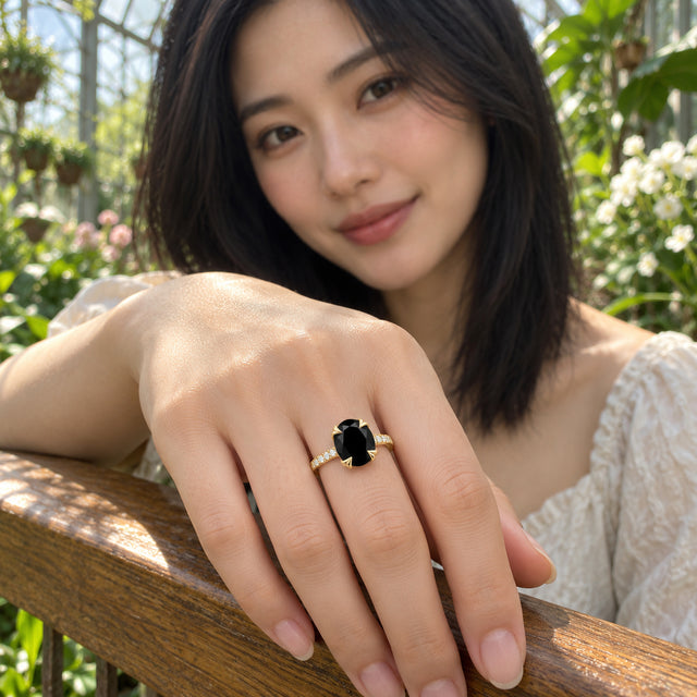 The yellow gold ring with an oval black stone is worn on a hand resting on a wooden railing among green plants.