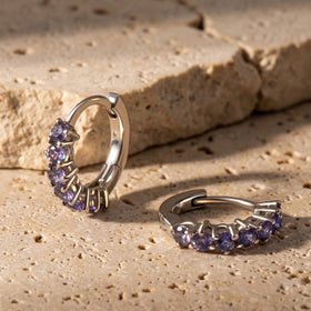 Two silver-tone violet gemstone hoops rest on a textured beige stone surface.