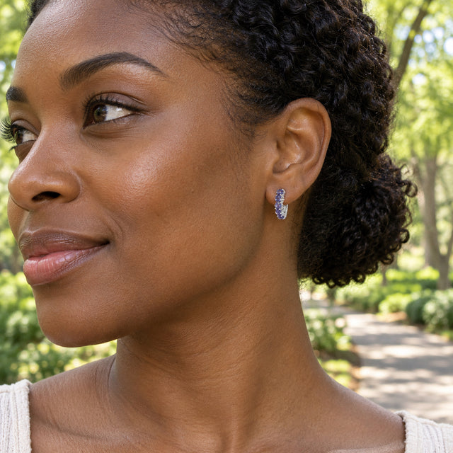 A woman with dark curly hair wears a small silver-tone violet gemstone hoop beside a leafy park path.
