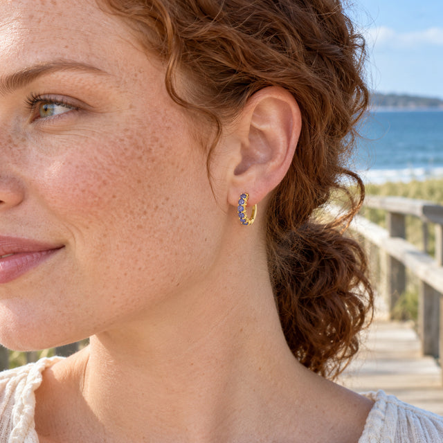 A freckled woman with auburn hair wears a yellow gold-tone violet gemstone hoop by a seaside boardwalk.
