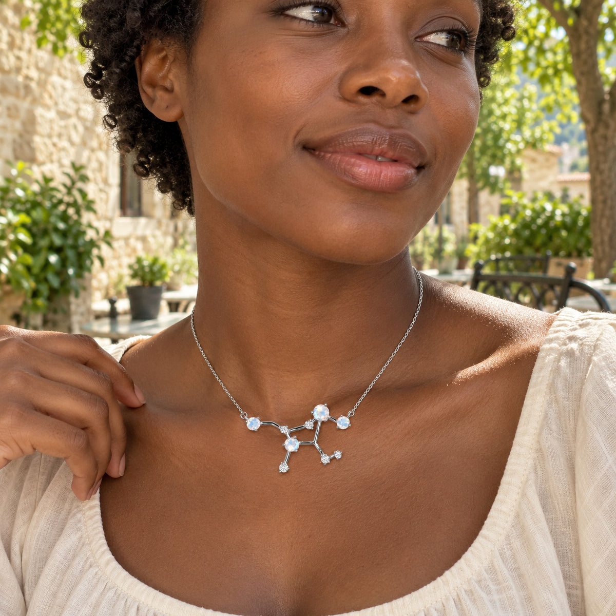 A woman with short curls wears a silver constellation necklace and a cream blouse in a sunlit stone courtyard.