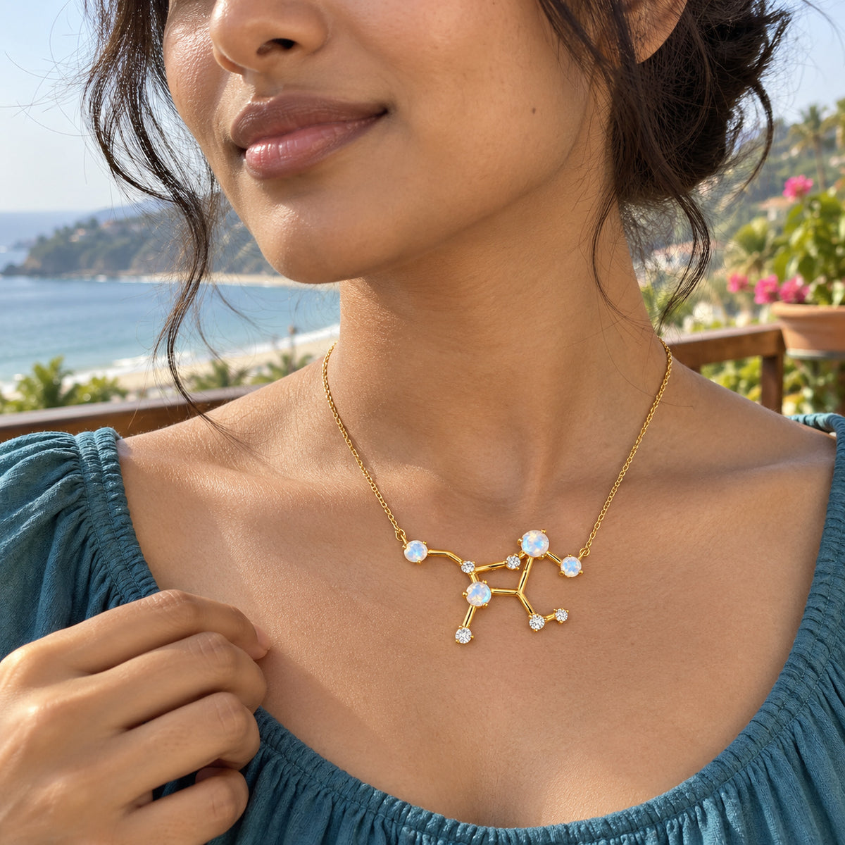 A woman with dark hair in a loose bun wears a yellow gold constellation necklace and a teal blouse on a seaside terrace.