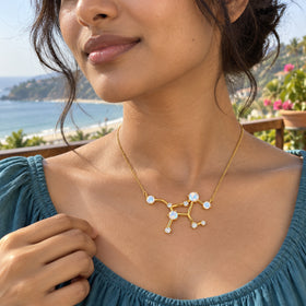 A woman with dark hair in a loose bun wears a yellow gold constellation necklace and a teal blouse on a seaside terrace.