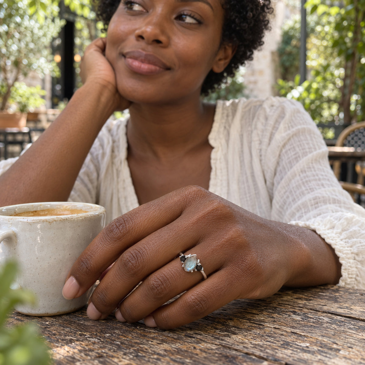 A sterling silver ring with a blue-gray pear-shaped stone is worn on a hand beside a ceramic cup in a leafy cafe.