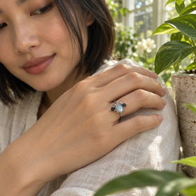 The silver ring is shown on a hand resting on a cream sleeve beside green plants and a stoneware planter.