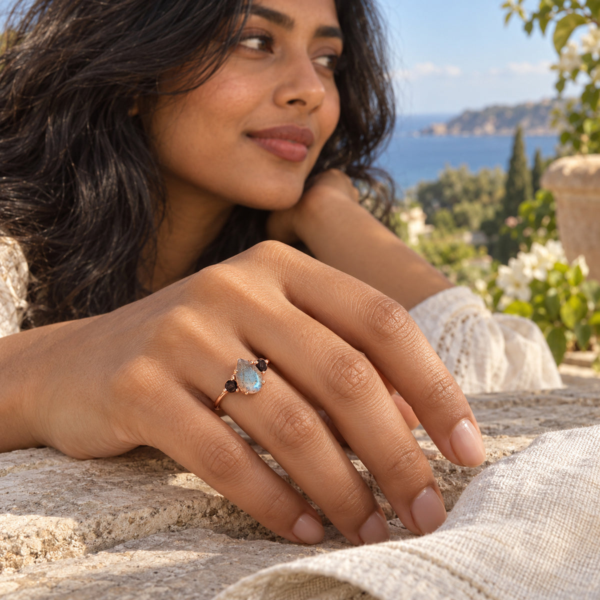 A rose gold vermeil ring with two black side stones is worn on a hand resting on a stone terrace overlooking the coast.