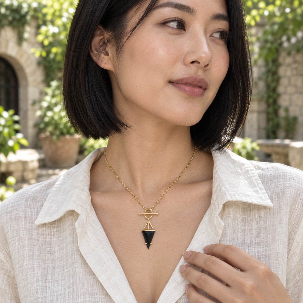 A woman in an ivory linen blouse wears a gold necklace with a black triangular pendant in a leafy courtyard.