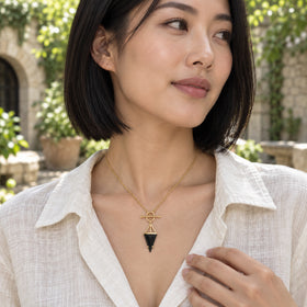 A woman in an ivory linen blouse wears a gold necklace with a black triangular pendant in a leafy courtyard.
