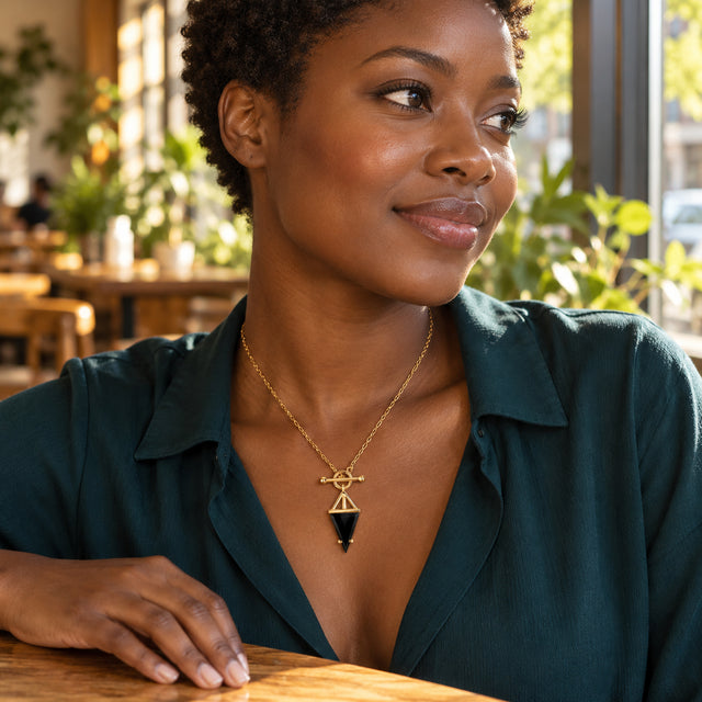 A woman in a dark teal blouse wears the black triangular pendant necklace beside a sunlit cafe window.